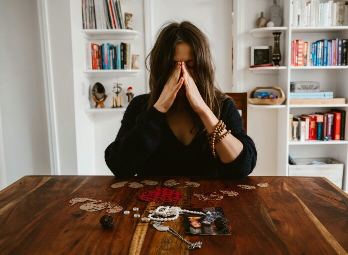 A woman in deep thought sits at a table with beads and a photo, conveying introspection. - emergency love spell for broken heart healing fast